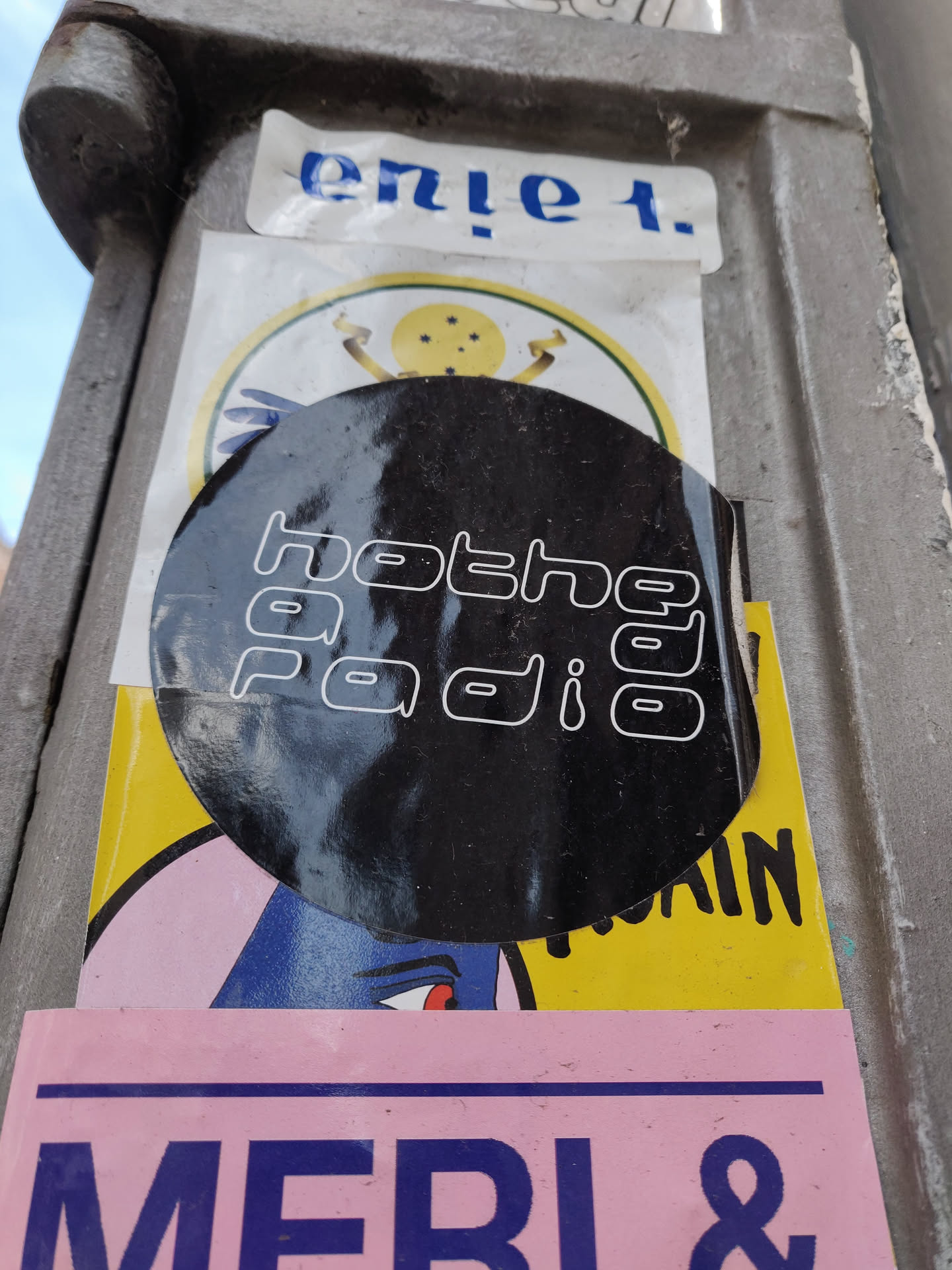 Black round sticker with the text "hotheya radio" in a stylized font.  It's stuck on a pole alongside other stickers.