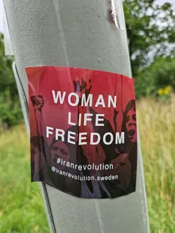 A sticker expressing solidarity with the Iranian Revolution.  It features the slogan "Woman Life Freedom" and includes a social media handle.