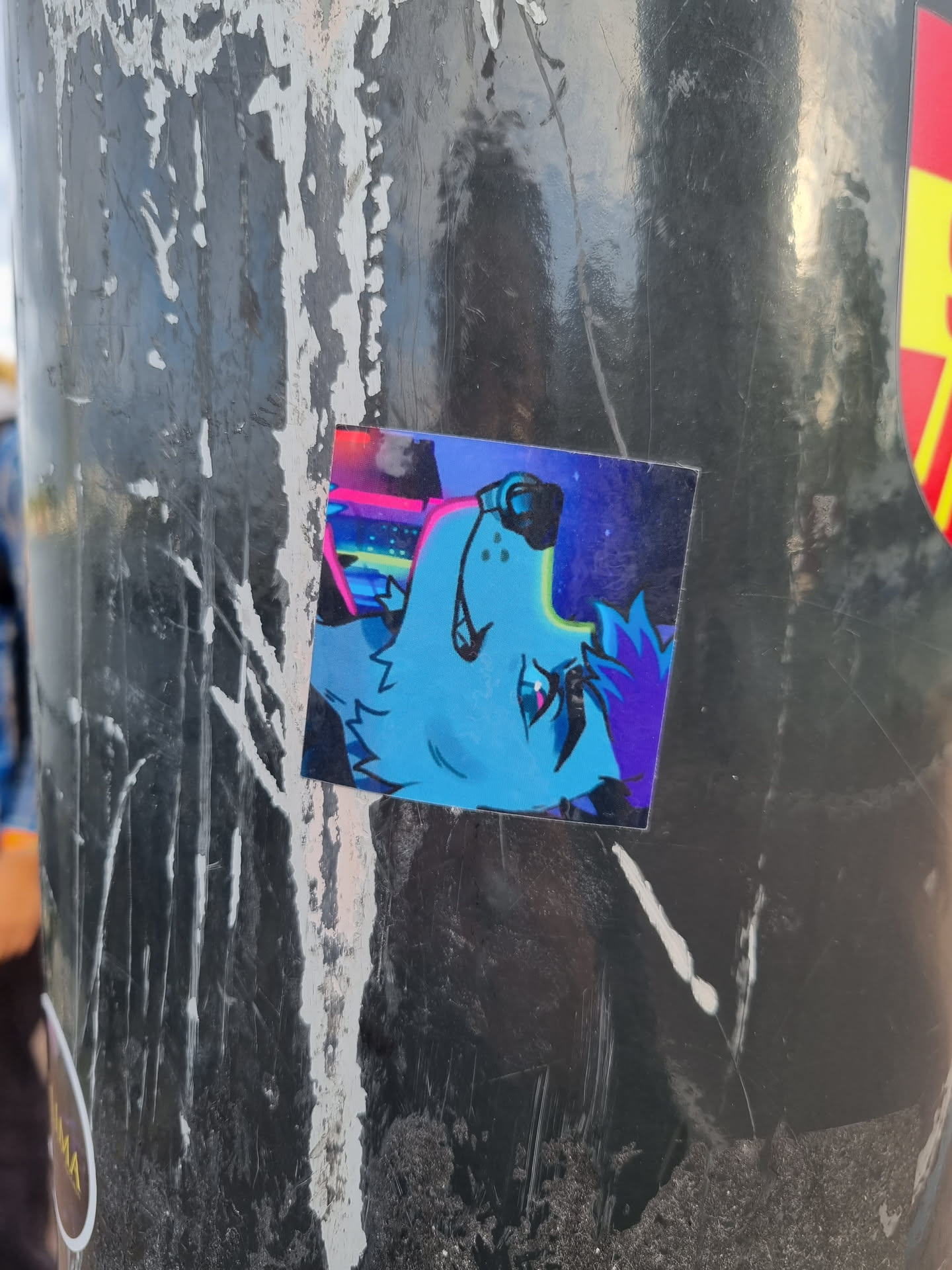 A vibrant sticker featuring a stylized creature with headphones, set against a neon-lit cityscape backdrop. The art style is reminiscent of cyberpunk and digital art, with a mix of bright colors and dark shadows. The sticker is affixed to a dark gray surface, which appears to be a metal pole.