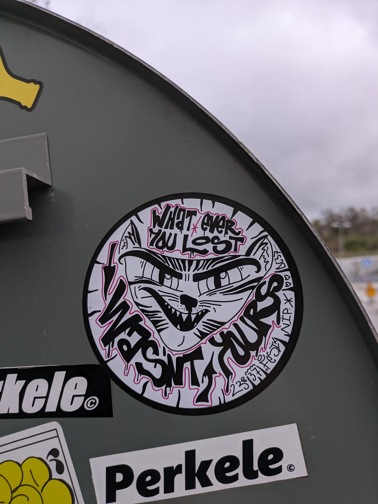 A round sticker features a stylized drawing of a cat's face with sharp teeth and intense eyes. The text "WHATEVER YOU LOST IT WASN'T YOURS" surrounds the cat's face in a dripping, graffiti-like font. Some additional symbols and characters are included around the edges of the sticker. The sticker is black and white with a subtle pink outline on the text.