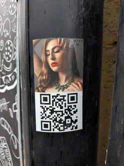 The sticker features a glossy, high-quality photograph of a woman with long, wavy brown hair, her eyes closed in a serene expression. She wears vibrant red lipstick and matching red nail polish on her visible hand, which is gently placed near her face. A prominent, intricate necklace made of green gemstones adorns her neck. The background of the portrait is dark and subtly textured. Below the portrait, a standard black and white QR code is printed on a white background, occupying the lower half of the sticker. The entire sticker is affixed to a dark, textured pole, showing some minor wear and tear around its edges.