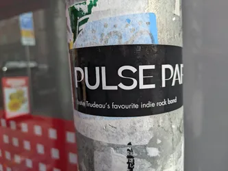 A black and white sticker with the text "PULSE PARTY" in a large, sans-serif font. Below the band name, it says "Justin Trudeau's favourite indie rock band". The sticker is affixed to a metal pole with other stickers surrounding it.  The sticker shows some signs of wear.