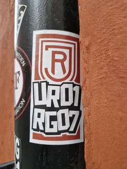 A brown and white sticker with the letters RJ in a logo, and the numbers UR01 RG07 underneath. The sticker is on a black pole.  The style is reminiscent of graffiti or street art.