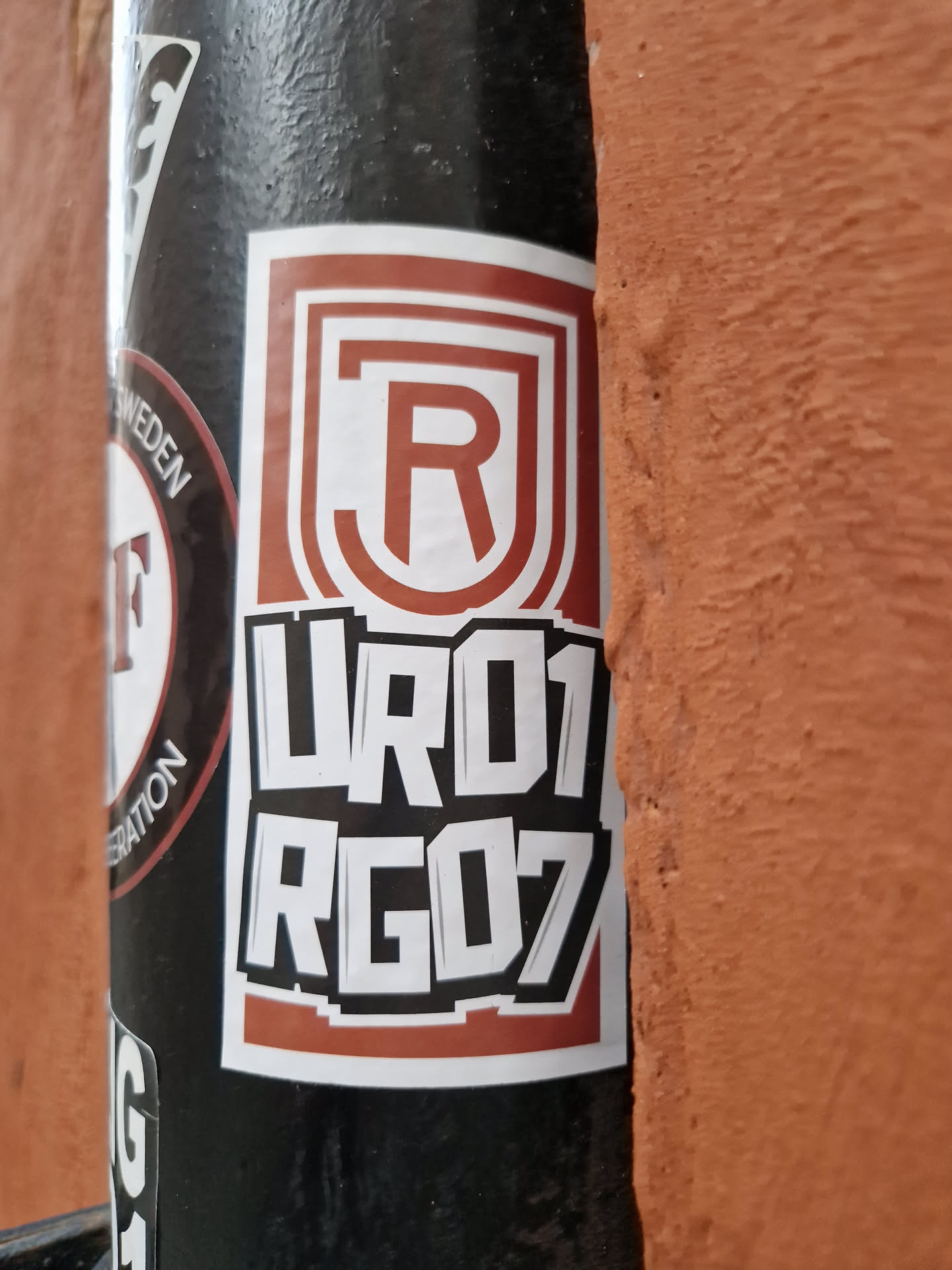 A brown and white sticker with the letters RJ in a logo, and the numbers UR01 RG07 underneath. The sticker is on a black pole.  The style is reminiscent of graffiti or street art.