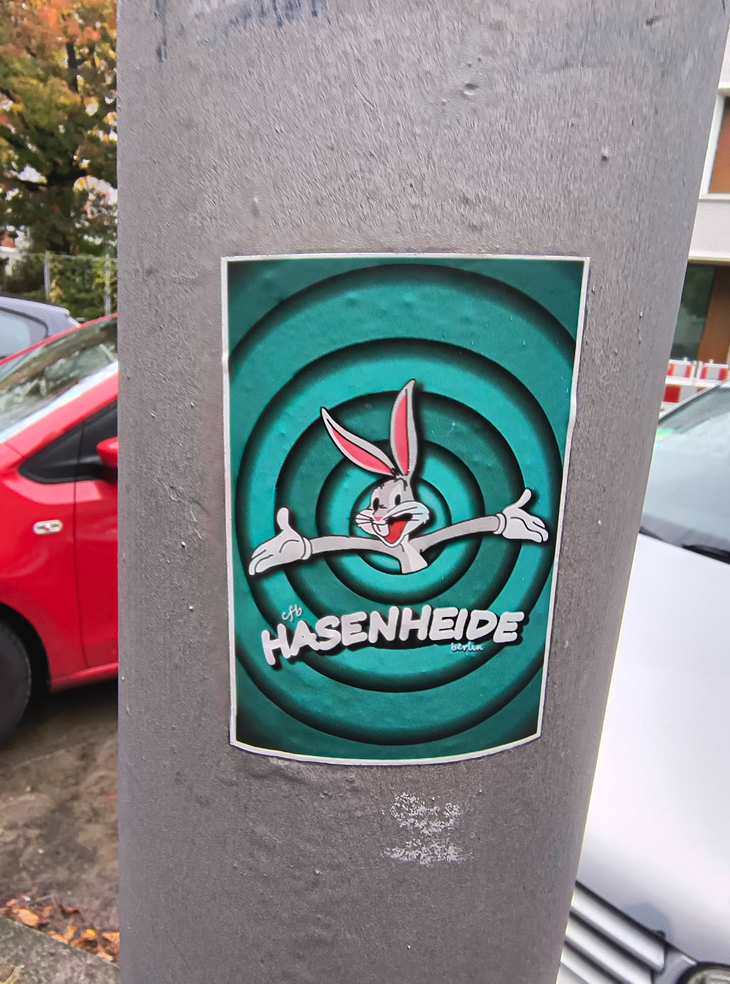 The sticker features a stylized depiction of Bugs Bunny, the iconic cartoon rabbit, against a vibrant teal background with concentric circles that create a sense of depth. Bugs Bunny is shown with his characteristic mischievous grin, arms outstretched in a welcoming gesture. He is rendered in shades of gray with pink inner ears and white gloves. Below the character, the word 'HASENHEIDE' is prominently displayed in bold, white, 3D-style lettering, with a smaller word 'berlin' underneath in a lighter font. The overall aesthetic is reminiscent of classic animation title cards.