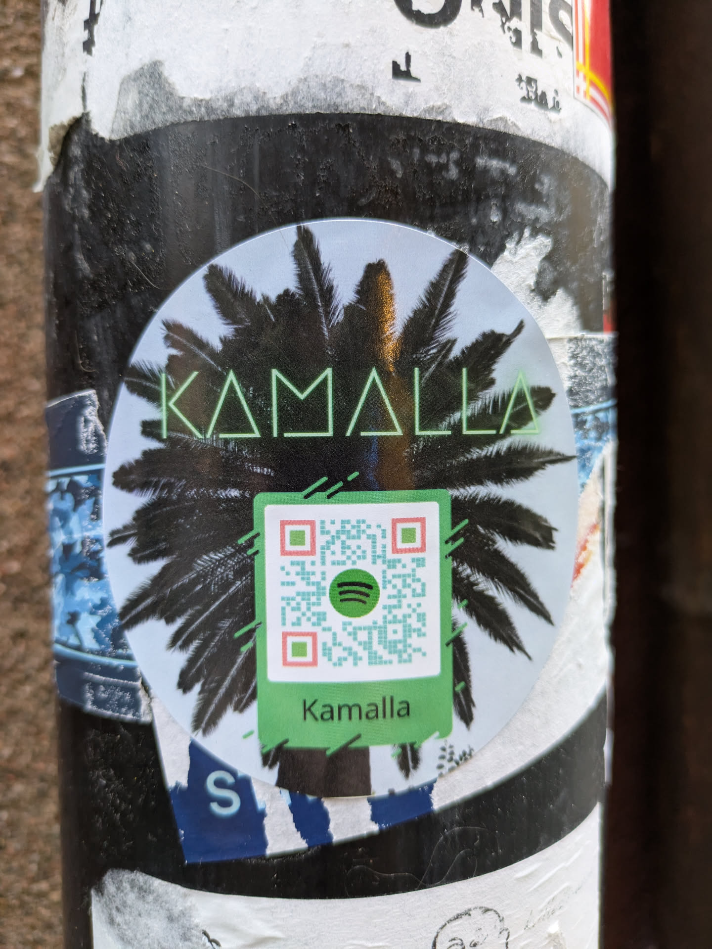 Round sticker featuring the text "Kamalla" in a stylized font, overlaid on an image of a palm tree. A QR code linking to Spotify is also present. 