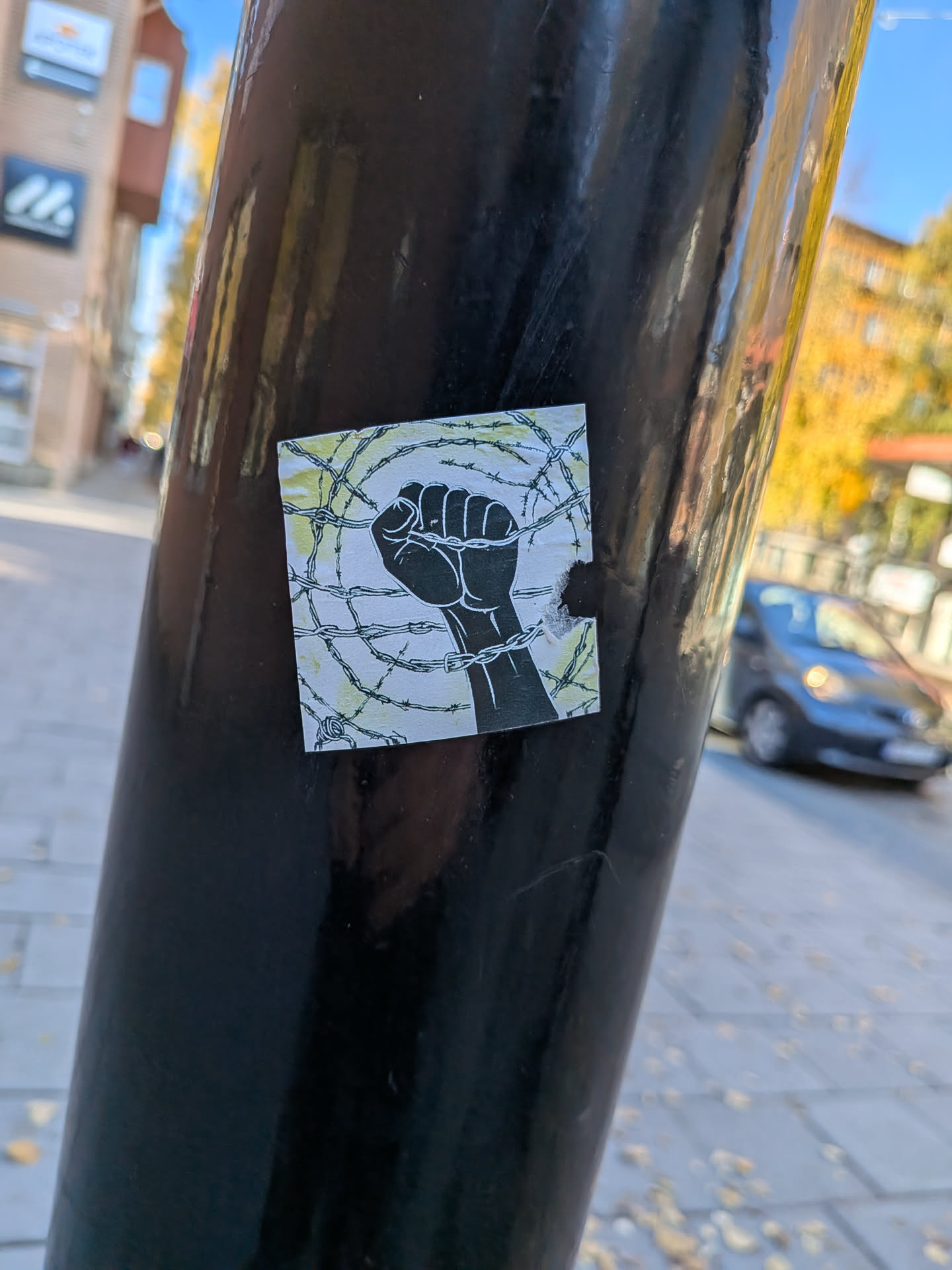 The sticker features a black silhouette of a clenched fist raised upwards, breaking through a tangled mess of barbed wire. The background is a light, textured yellow with subtle green lines suggesting more barbed wire or a distressed texture. The overall impression is one of struggle and resistance.