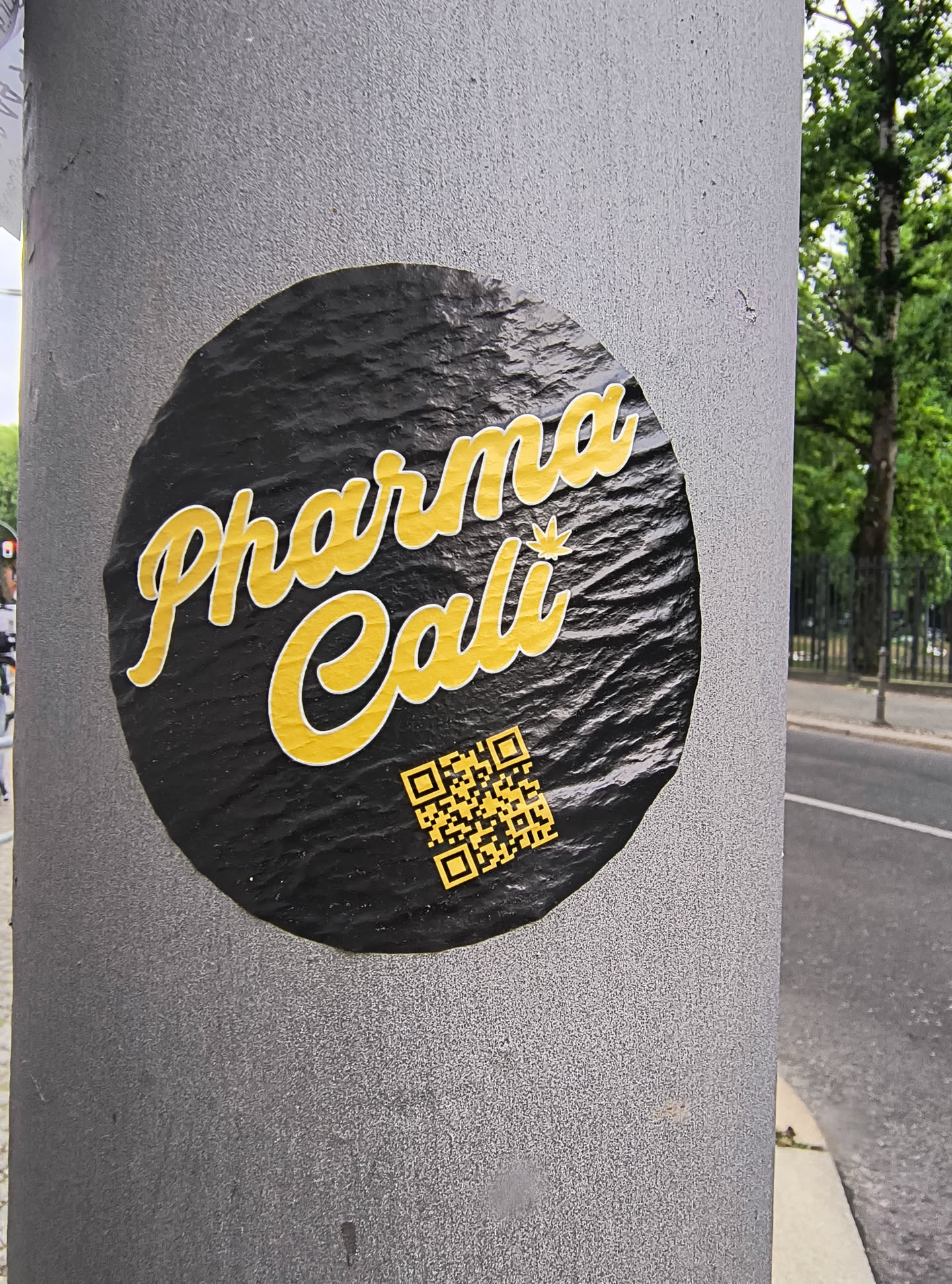 This black and yellow round sticker affixed to a pole features the text 'Pharma Cali' in a stylized font. Below the text, a QR code is present, presumably leading to more information about Pharma Cali.