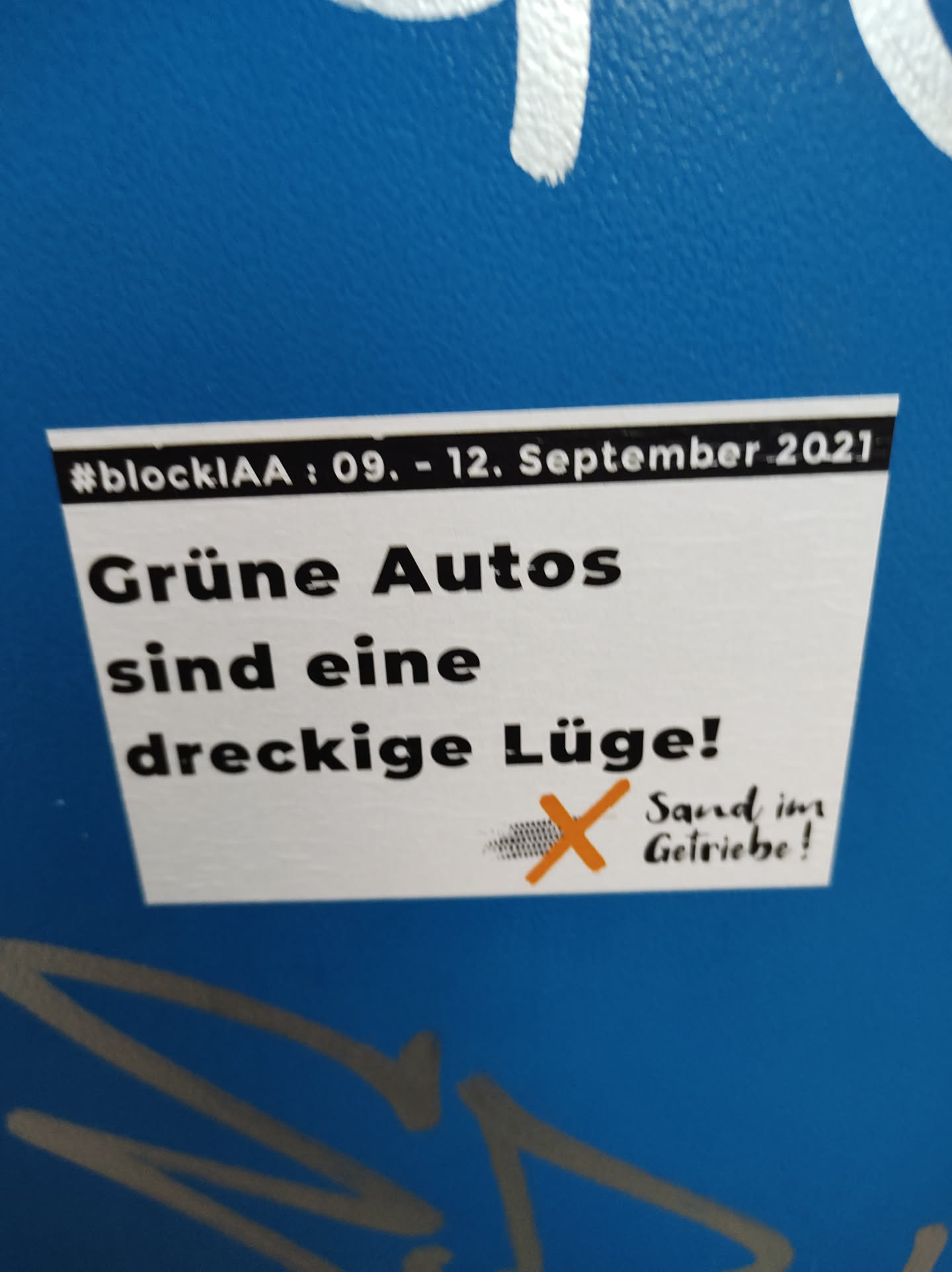 Sticker expressing a critical opinion on green cars, possibly referencing the event #blockIAA from September 9-12, 2021.  It features a crossed-out image suggesting sabotage. Text says: "Green cars are a dirty lie! Sand in the gears!"