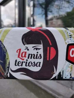 Sticker featuring a stylized illustration of a girl with headphones, with the text "La misteriosa" underneath. The sticker has a predominantly dark color scheme with accents of red. 