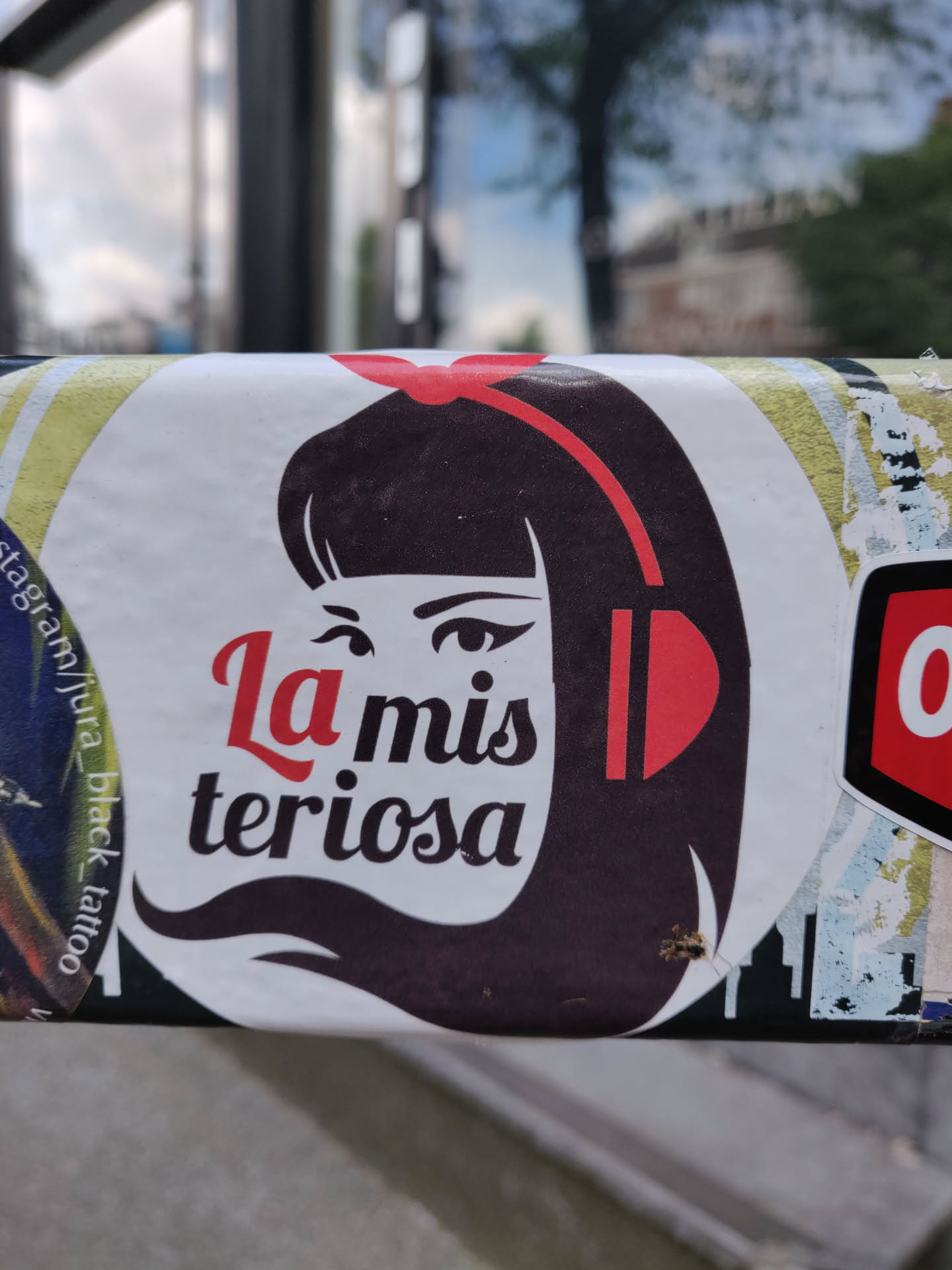 Sticker featuring a stylized illustration of a girl with headphones, with the text "La misteriosa" underneath. The sticker has a predominantly dark color scheme with accents of red. 