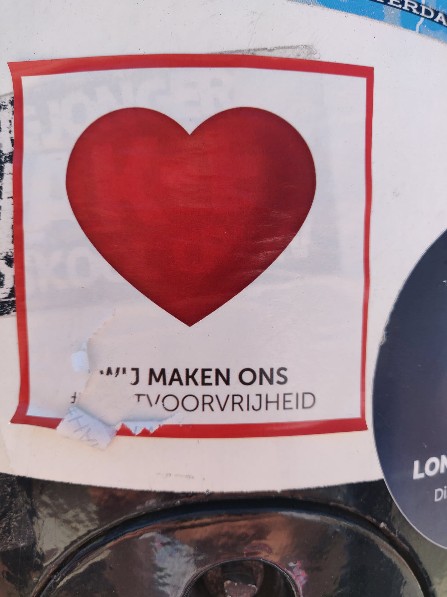 A white square sticker with a red heart in the center and a phrase in Dutch at the bottom. The phrase seems to be incomplete. 