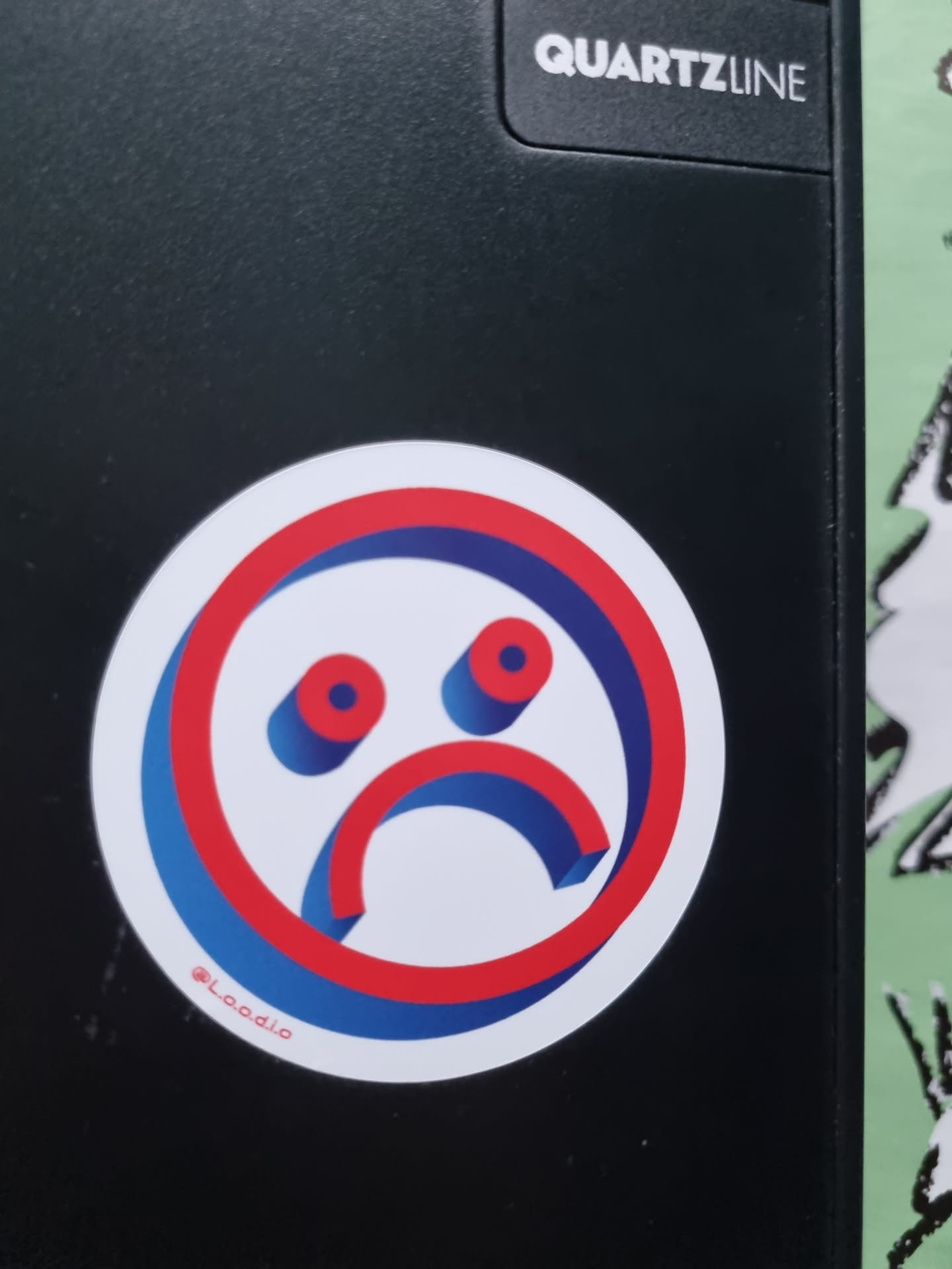 A round sticker with a stylized sad face in red, white and blue. The face is designed in a 3D style with a thick border. The sticker is placed on a black surface.  It includes the social media handle @Laadio.