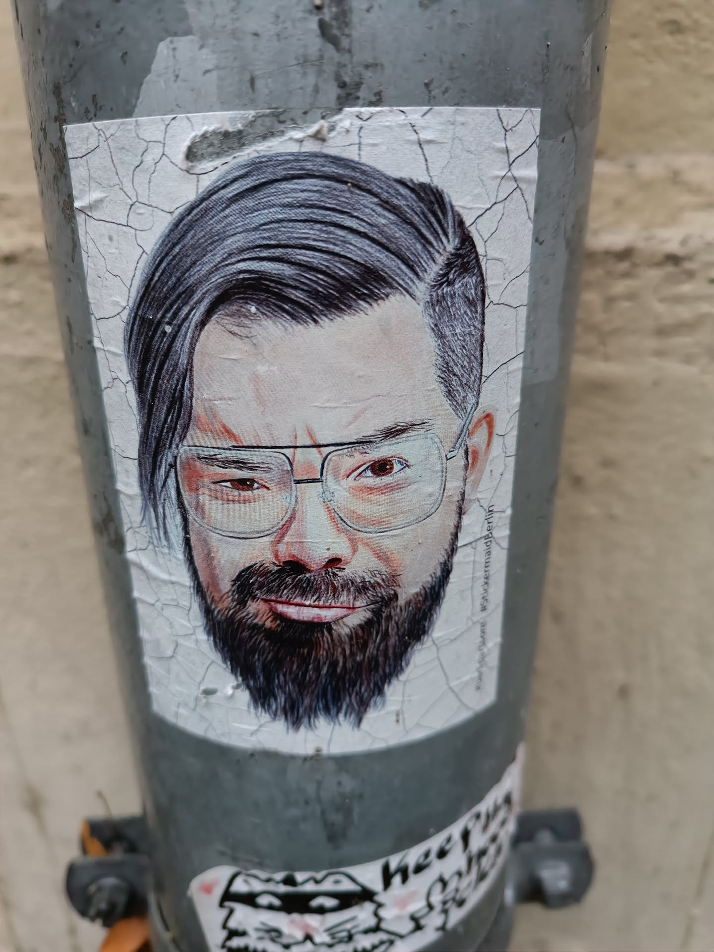 Close-up shot of a sticker featuring a realistic pencil sketch of a man's face. The man has dark hair styled neatly, is wearing glasses, and has a well-groomed beard. The sticker is affixed to a weathered gray pole, showing some peeling around the edges.
