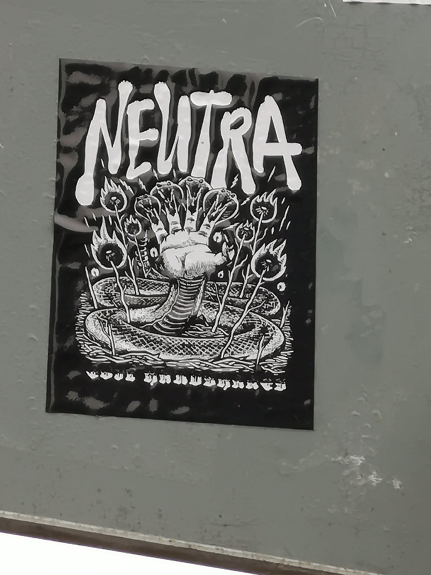 The sticker is a rectangular, black and white design. At the top, the word "NEUTRA" is prominently displayed in a bold, distressed white font against a black background. The central illustration features a large, coiled snake with multiple heads, from which a human-like fist emerges. The snake's scales are intricately detailed. Surrounding the snake and fist are several thin, plant-like stalks, each topped with a stylized flame or burning orb. The artwork has a strong, high-contrast, illustrative style, reminiscent of woodcut or linocut prints, with white lines defining elements against a dark, textured background. There is a small, unreadable line of text or symbols at the very bottom.