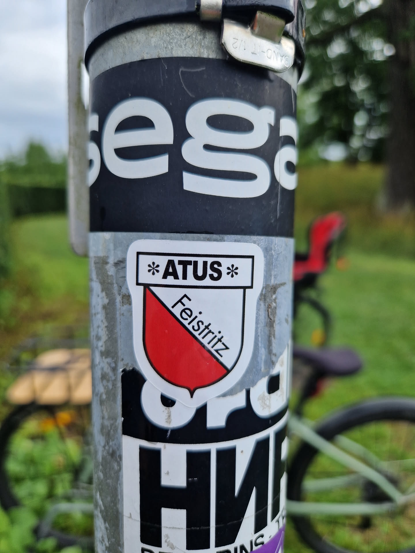 A close-up photo of a sticker featuring the logo of ATUS Feistritz am See. The sticker is affixed to a metal pole, with other stickers visible in the background.  The main sticker is shield-shaped, with a red and white design and the text "Feistritz" written on it.