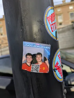 The sticker features a brightly lit image of two young men, both smiling and pointing upwards with their left hands. They are wearing orange t-shirts with a red collar and a white Nike swoosh on the left chest, suggestive of sports apparel. Behind them, a dartboard is partially visible, indicating a casual or recreational setting. A QR code is placed prominently on the lower left side of the sticker. The top of the sticker has a blue gradient with text in white and pink lettering.
