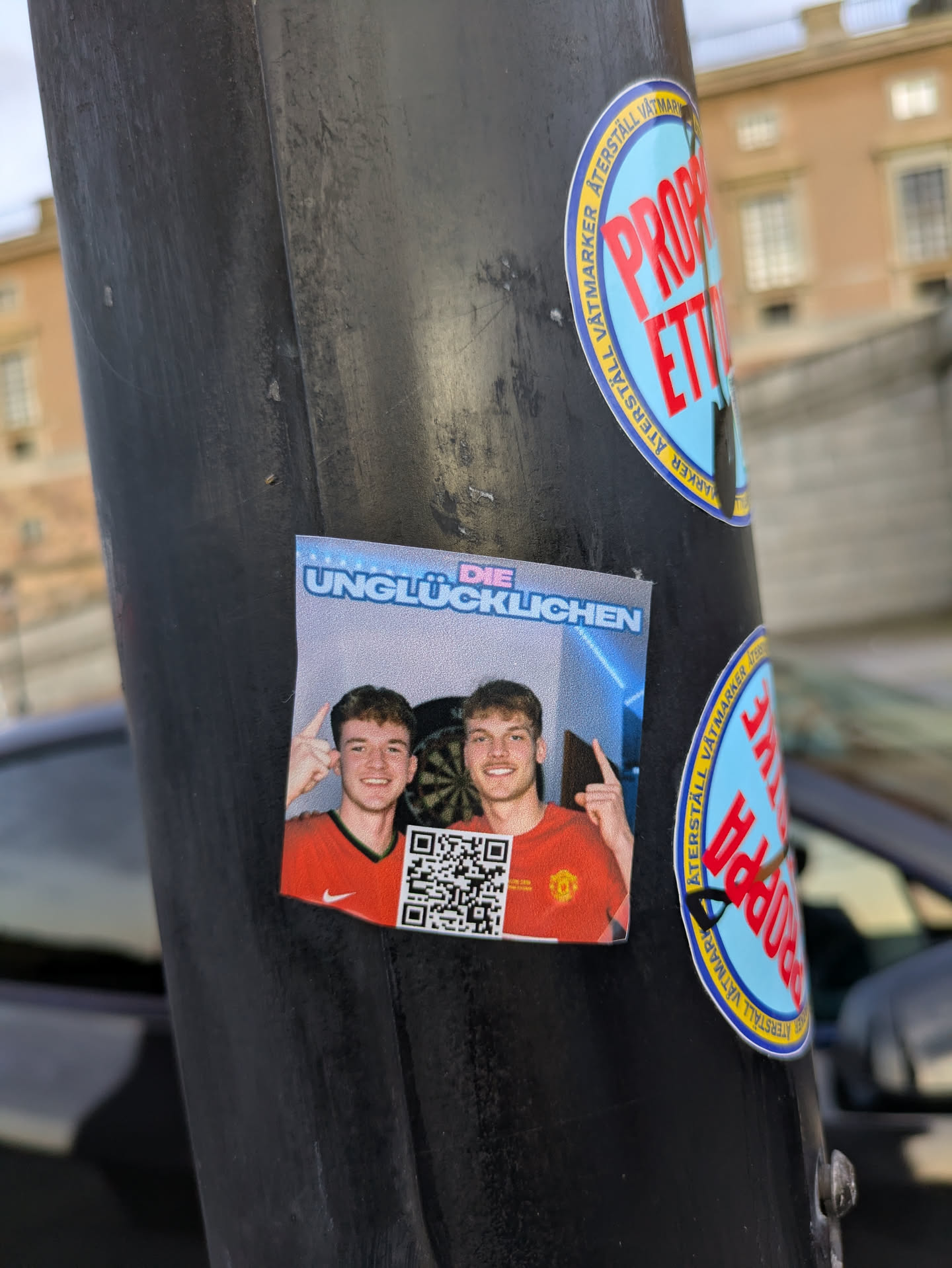 The sticker features a brightly lit image of two young men, both smiling and pointing upwards with their left hands. They are wearing orange t-shirts with a red collar and a white Nike swoosh on the left chest, suggestive of sports apparel. Behind them, a dartboard is partially visible, indicating a casual or recreational setting. A QR code is placed prominently on the lower left side of the sticker. The top of the sticker has a blue gradient with text in white and pink lettering.