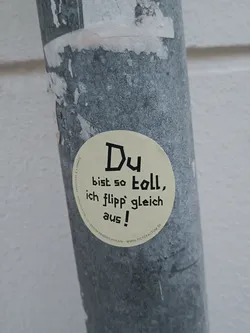 A round beige sticker with black text on a gray metal pole.  The text says "Du bist so toll, ich flipp gleich aus!" which translates from German to "You are so great, I'm about to flip out!"