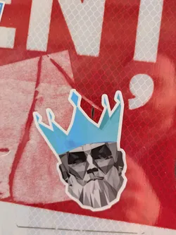 A sticker featuring a geometric representation of a man's head wearing a blue crown is affixed to a red and white surface. The design is stylized with a mosaic of gray and white polygons to form the head and beard. The crown is a bright blue and also constructed with geometric shapes.