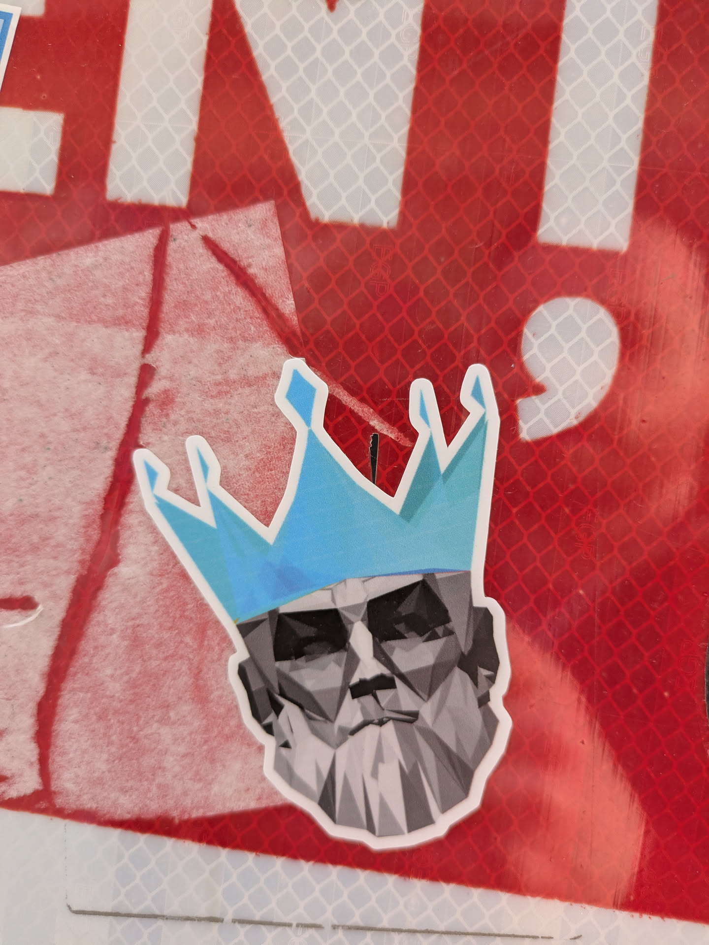 A sticker featuring a geometric representation of a man's head wearing a blue crown is affixed to a red and white surface. The design is stylized with a mosaic of gray and white polygons to form the head and beard. The crown is a bright blue and also constructed with geometric shapes.
