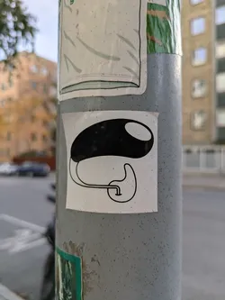 A minimalistic sticker featuring a black bean-like shape with a small tube attached to its bottom. It's affixed to a grey pole, surrounded by other stickers.
