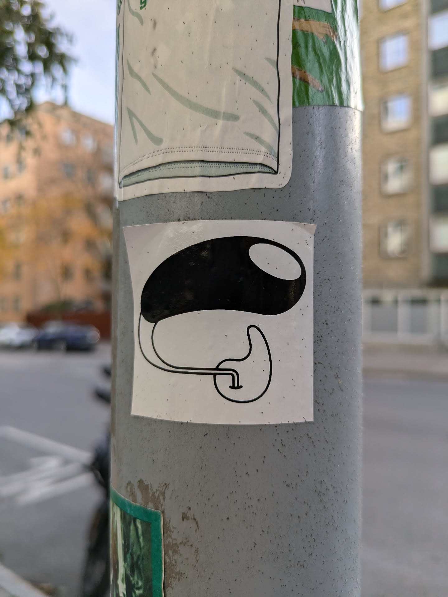 A minimalistic sticker featuring a black bean-like shape with a small tube attached to its bottom. It's affixed to a grey pole, surrounded by other stickers.