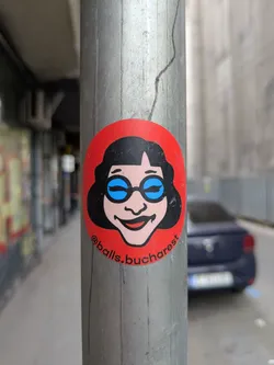 A round sticker on a metal pole shows a cartoon woman with black hair, blue glasses, and a smile. The sticker has a red background and the text '@balls.bucharest' is printed on it.