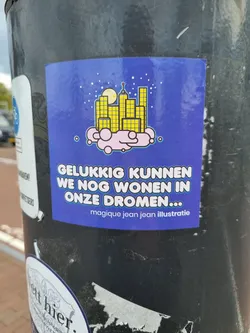 A sticker with an illustration of a city on a cloud at night. The text on the sticker says 'Gelukkig kunnen we nog wonen in onze dromen...' which translates to 'Luckily we can still live in our dreams...' in English.  The sticker also credits 'magique jean jean illustratie'.