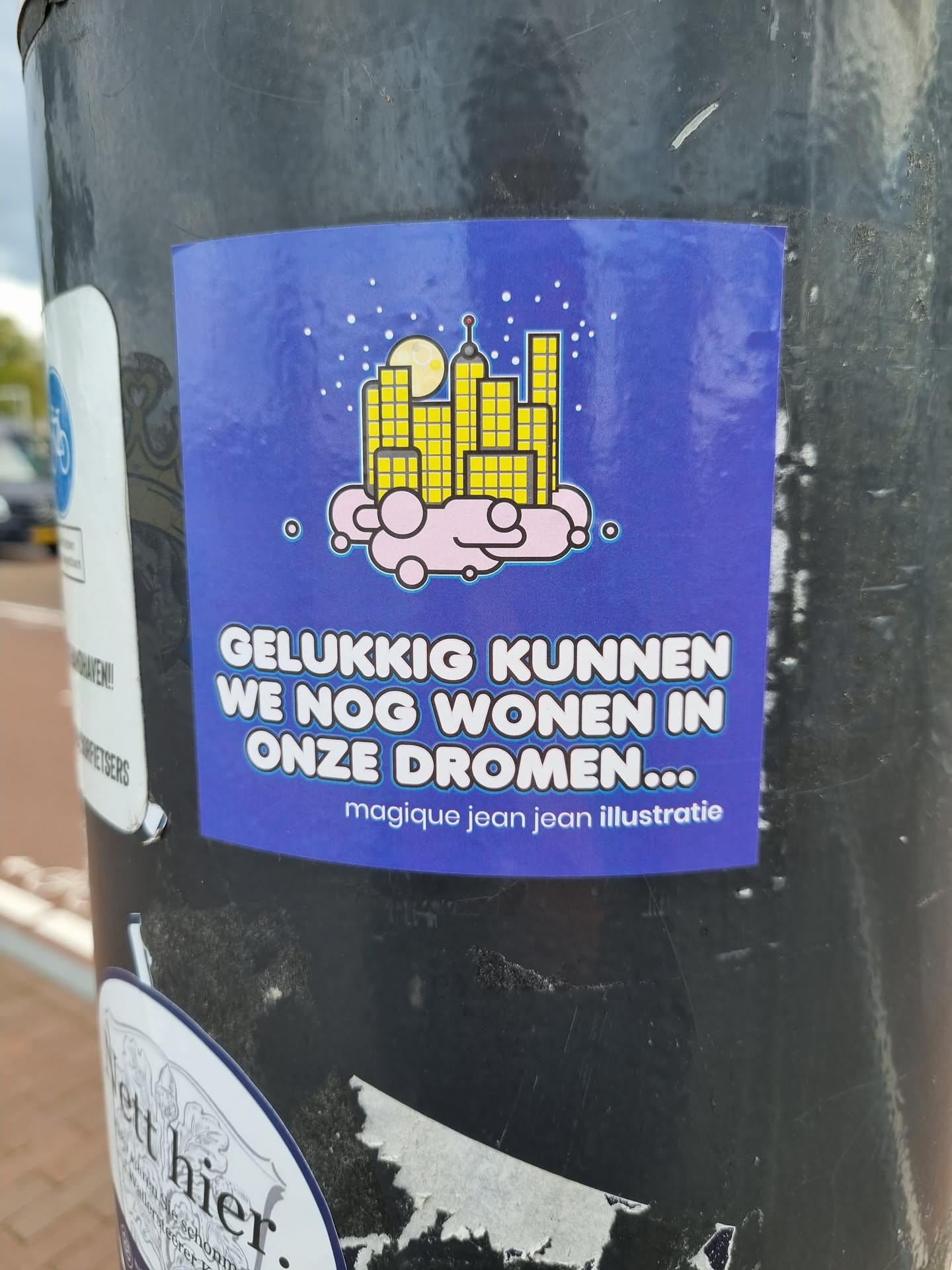 A sticker with an illustration of a city on a cloud at night. The text on the sticker says 'Gelukkig kunnen we nog wonen in onze dromen...' which translates to 'Luckily we can still live in our dreams...' in English.  The sticker also credits 'magique jean jean illustratie'.