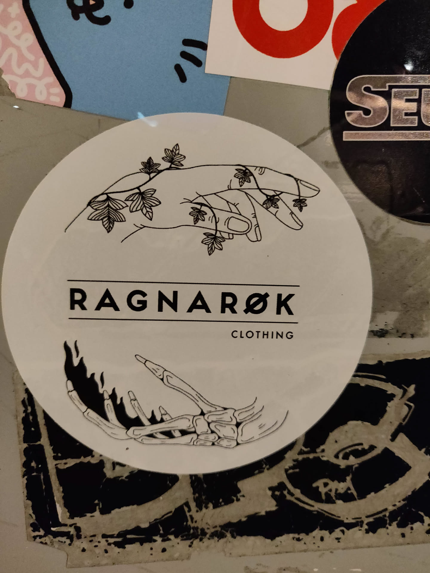 Round white sticker with black line art depicting a hand intertwined with leaves on one side and a skeleton hand engulfed in flames on the other.  The text "RAGNAROK" and "CLOTHING" is visible in the center.