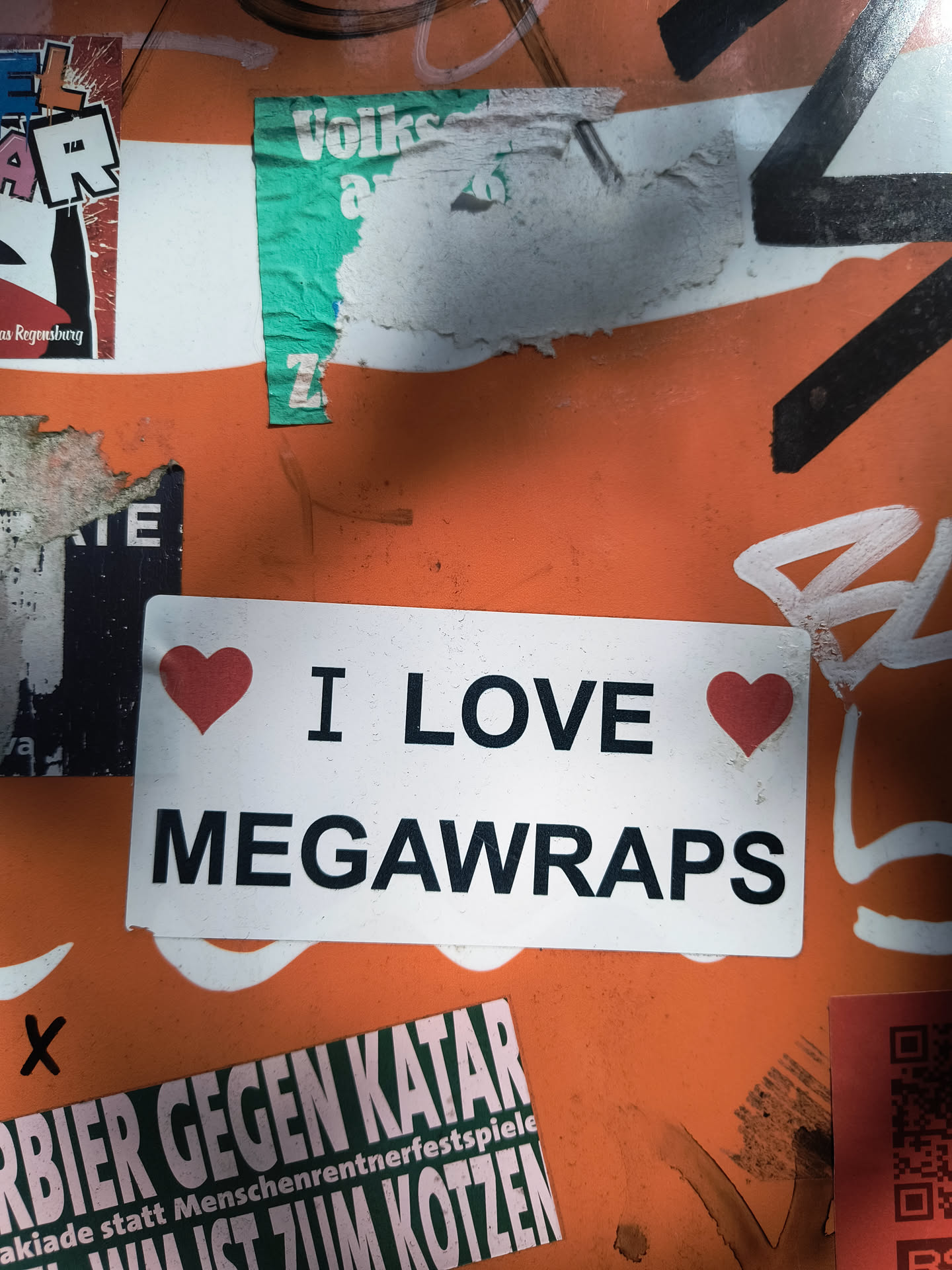 A rectangular white sticker featuring bold black sans-serif text. The phrase 'I LOVE MEGAWRAPS' is displayed, with two small red heart symbols flanking the word 'LOVE' – one before it and one after. The sticker has a clean, minimalist design and appears slightly worn or dirty on its surface, suggesting it is affixed to a public wall or object amidst other graffiti and stickers.