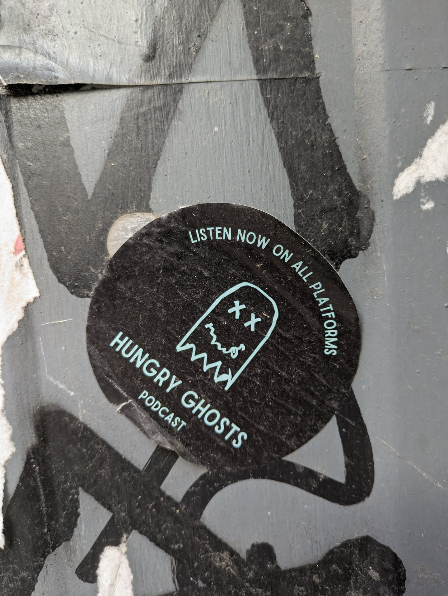 The sticker is round and black with the text "LISTEN NOW ON ALL PLATFORMS" around the edge. It features a teal ghost illustration with "XX" eyes and a wavy mouth. The text "HUNGRY GHOSTS" is printed below the ghost, with "PODCAST" underneath.