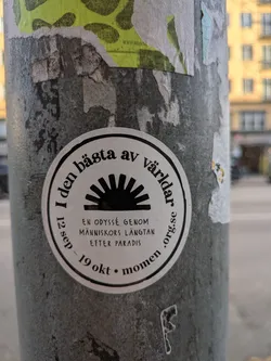 The sticker is circular with a black border. Inside, at the top, is text in a curved, serif font. Below this text is a black graphic of a sunburst or radiating light. Beneath the sunburst, there is more text in a smaller, sans-serif font, arranged in lines. The outer edge of the sticker also contains text, including dates and a website address.