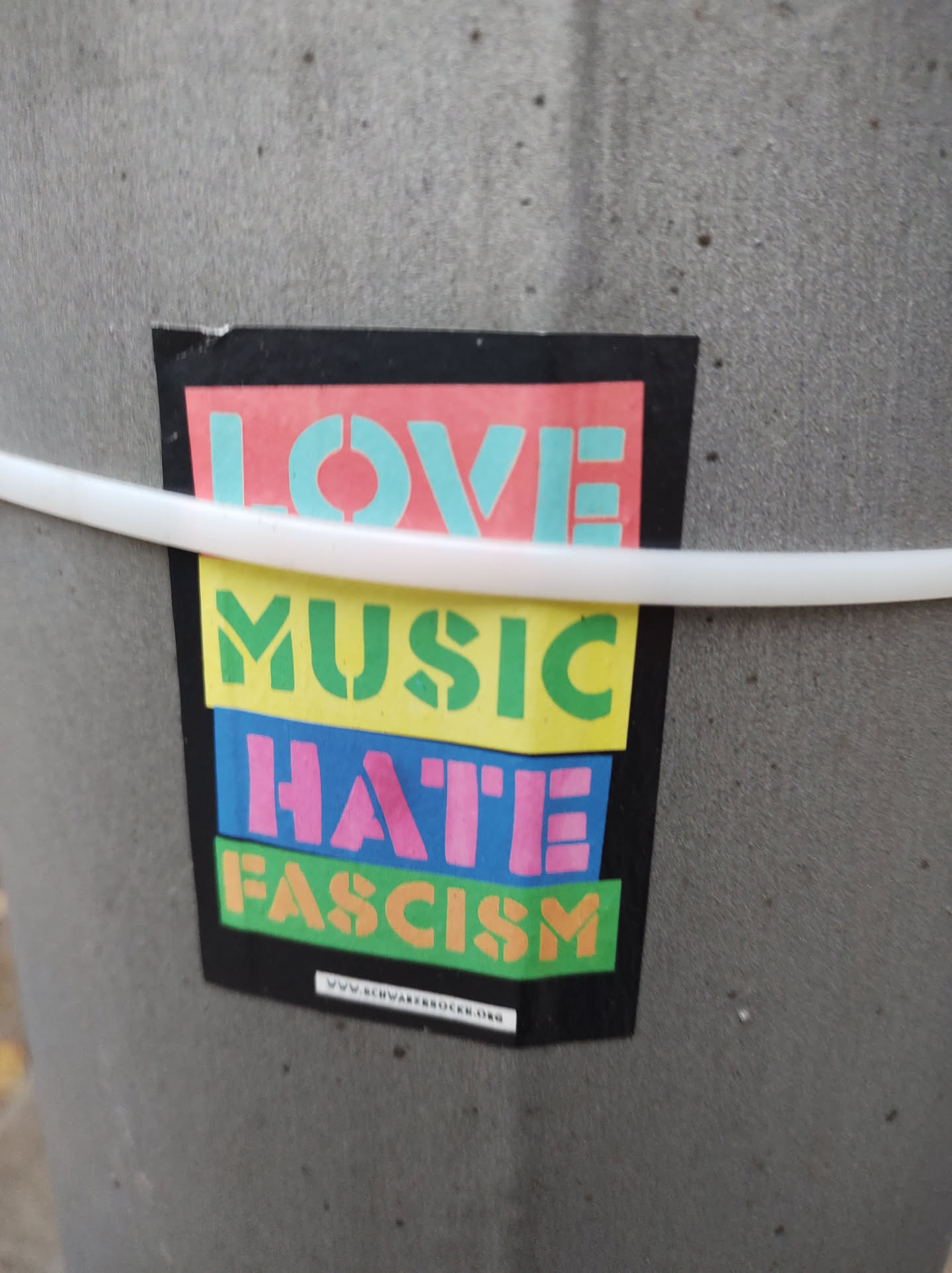 A sticker with the message 'Love Music Hate Fascism' in colorful block letters.  The sticker is affixed to a gray pole. A website address is visible at the bottom.