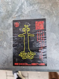 Black sticker with a yellow alien-like figure and a QR code. The text Excoliens is at the bottom of the sticker. Some illegible text is on the right side of the sticker.