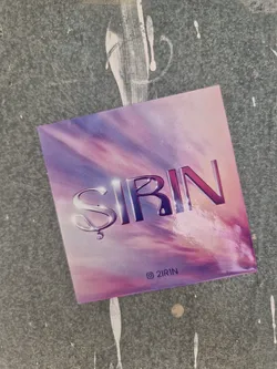 A sticker featuring the word SIRIN in a stylized font, set against a backdrop of purple and pink clouds. The sticker has a glossy finish and a minimalist design.  The artist's Instagram handle is visible in the bottom right corner.