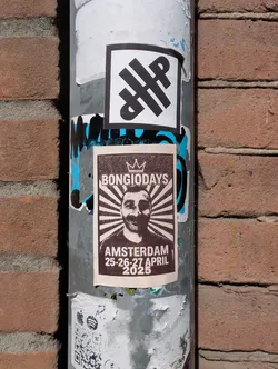 A poster on a pole advertises "Bongiodays" in Amsterdam from April 25-27, 2025. The poster features a portrait of a man with a mustache. There's another sticker with an abstract black design above the poster and a QR code sticker below it.