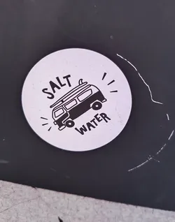 A round sticker featuring a black and white illustration of a van with a surfboard on top, surrounded by the text "SALT WATER". The sticker has a minimalist, hand-drawn style.