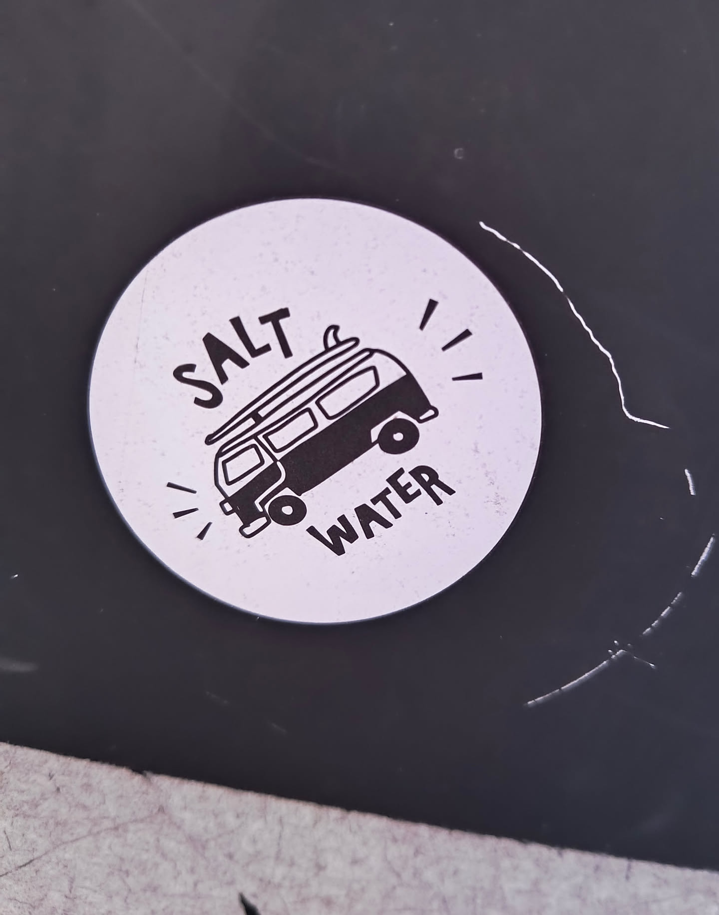 A round sticker featuring a black and white illustration of a van with a surfboard on top, surrounded by the text "SALT WATER". The sticker has a minimalist, hand-drawn style.