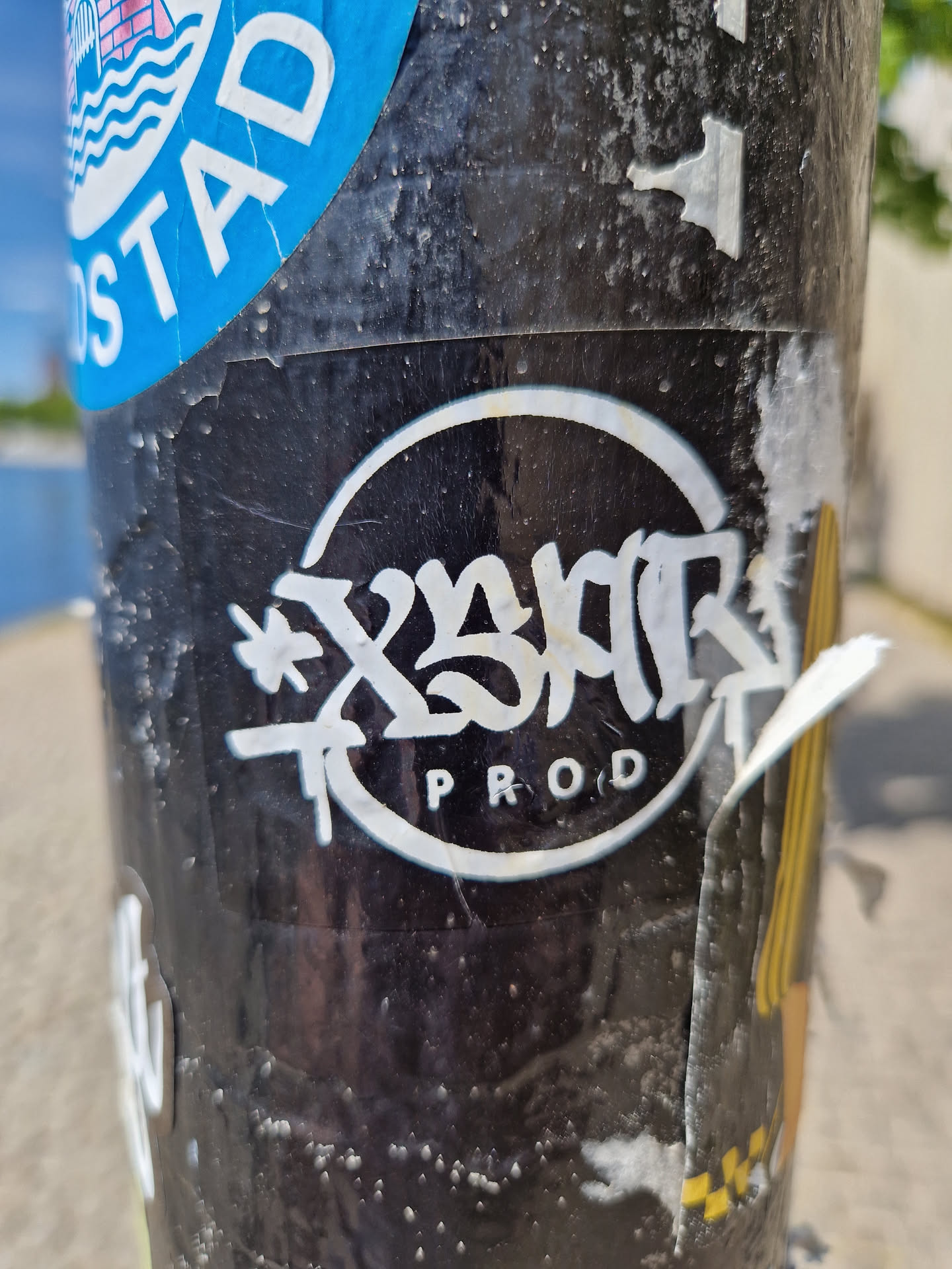 A black and white circular sticker with stylized graffiti text that says "X3MR Prod." The style is reminiscent of street art and is applied to a weathered surface. 