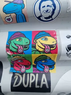 Street sticker This sticker features a pop art-style design with four colorful reptile characters, each wearing a beanie and sticking out their tongues. Each character is set in a different colored square, creating a vibrant, eye-catching design. The word "DUPLA" is prominently displayed below the characters. The style is reminiscent of 1960s pop art.
