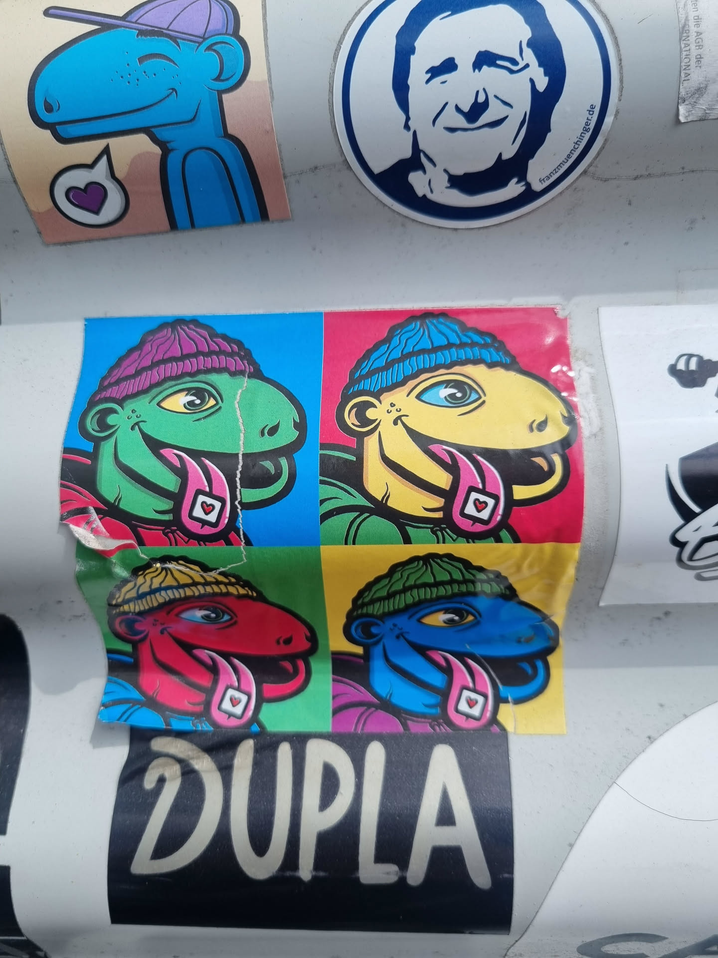 This sticker features a pop art-style design with four colorful reptile characters, each wearing a beanie and sticking out their tongues. Each character is set in a different colored square, creating a vibrant, eye-catching design. The word "DUPLA" is prominently displayed below the characters. The style is reminiscent of 1960s pop art.