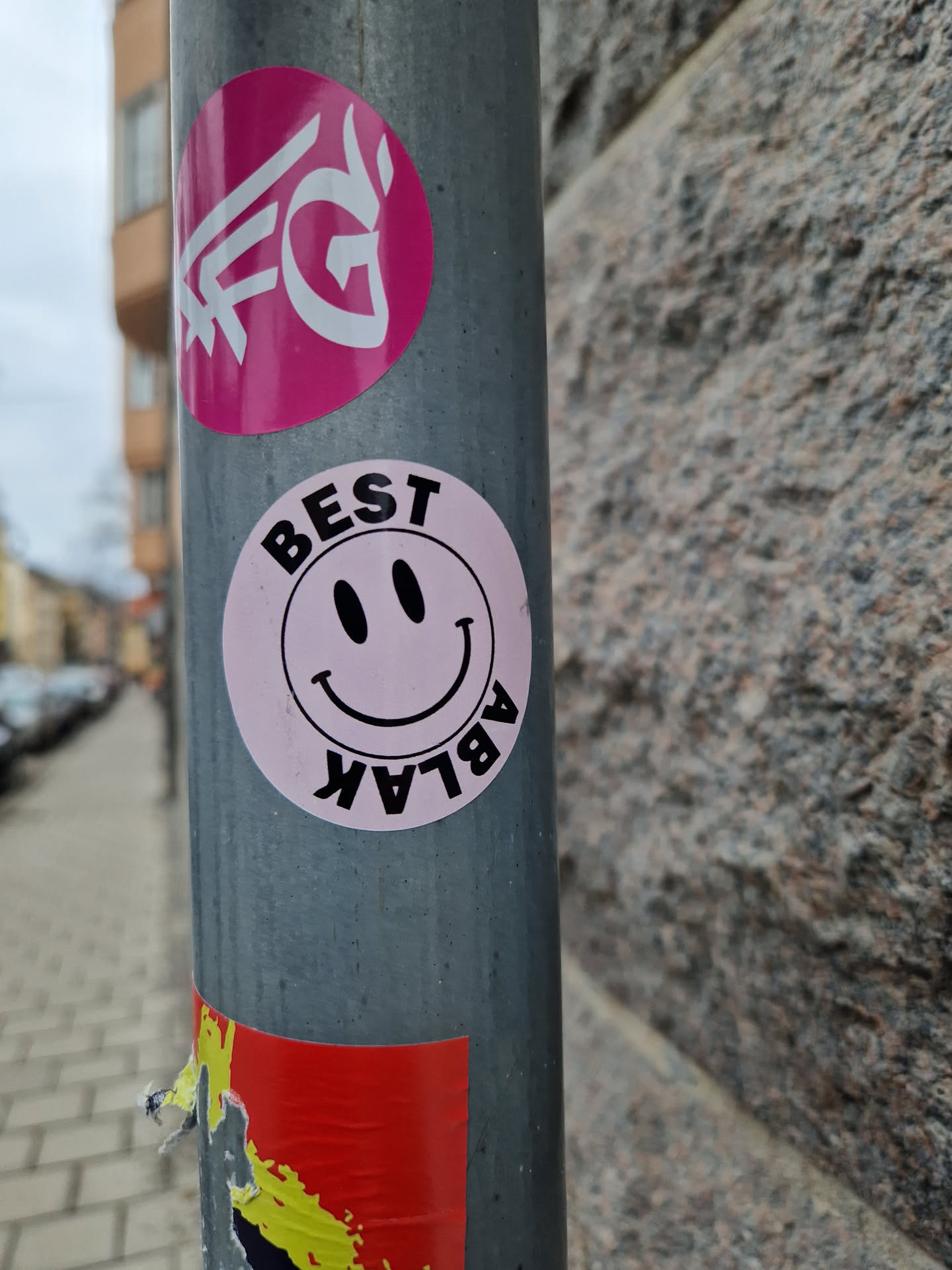 A pink and black round sticker with a simple smiley face and the word 'BEST' above it and 'ABLAK' written upside down below it. It's affixed to a grey metal pole alongside other stickers.