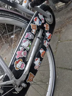 The image features a collection of numerous die-cut stickers applied to a black bicycle fender. The stickers predominantly showcase popular Sanrio characters, including Hello Kitty in various outfits and poses (e.g., in a pink dress, a blue outfit, a red outfit, holding a pumpkin, on a surfboard, in a green inner tube) and My Melody in a pink dress. Interspersed among these are several brown bear characters, resembling Rilakkuma, in simple, cute poses. Each sticker has a distinct white border, and they are arranged somewhat haphazardly across the fender. The color palette is vibrant, featuring white, pink, red, blue, green, brown, orange, and yellow, creating a cheerful and playful aesthetic.
