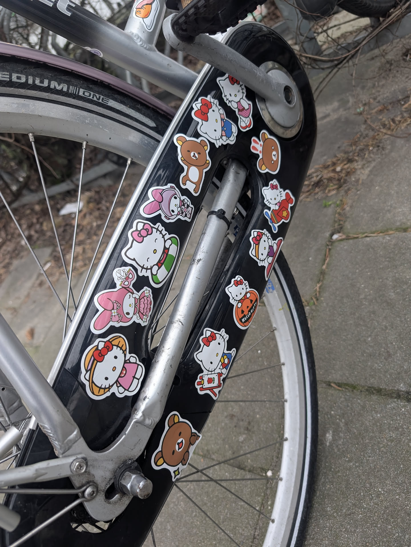 The image features a collection of numerous die-cut stickers applied to a black bicycle fender. The stickers predominantly showcase popular Sanrio characters, including Hello Kitty in various outfits and poses (e.g., in a pink dress, a blue outfit, a red outfit, holding a pumpkin, on a surfboard, in a green inner tube) and My Melody in a pink dress. Interspersed among these are several brown bear characters, resembling Rilakkuma, in simple, cute poses. Each sticker has a distinct white border, and they are arranged somewhat haphazardly across the fender. The color palette is vibrant, featuring white, pink, red, blue, green, brown, orange, and yellow, creating a cheerful and playful aesthetic.