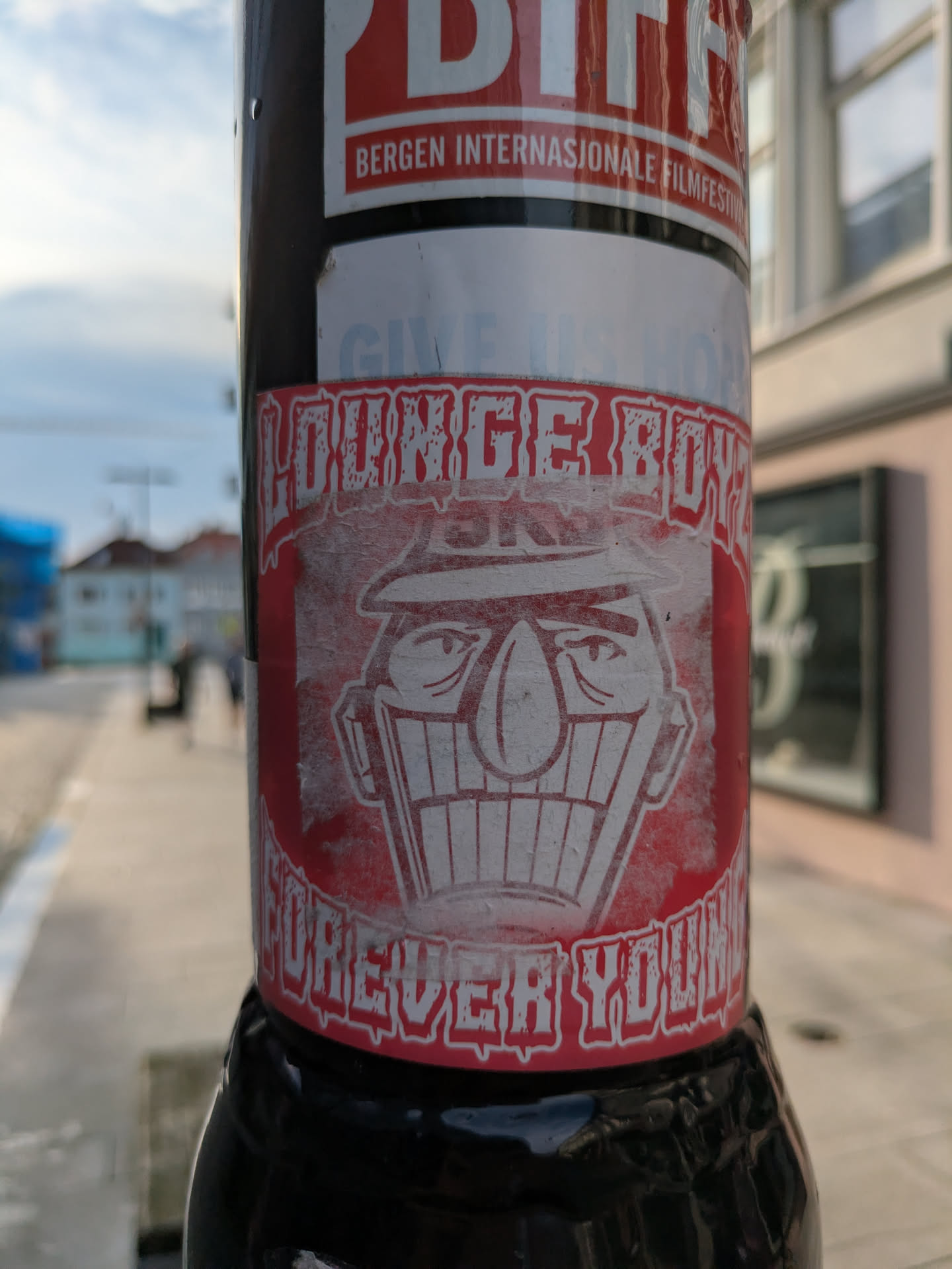 The central sticker is round, predominantly red with white outlines and text. It displays a distinctive, cartoonish character's face wearing a fedora-style hat, with a large, somewhat sinister grin revealing many teeth. The eyes are small and narrow, giving the character a shifty or mischievous look. The text 'LOUNGE BOYS' is arched above the character's head, and 'FOREVER YOUNG' is curved below, both in a distressed, blocky white font. The sticker shows signs of wear, with some fading and peeling.