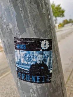 A sticker featuring a man's portrait with the text 'A105' and 'Fernet' underneath. A thought bubble with a bottle graphic is present above the man's head. The sticker shows some wear, suggesting it has been exposed to the elements for a while.