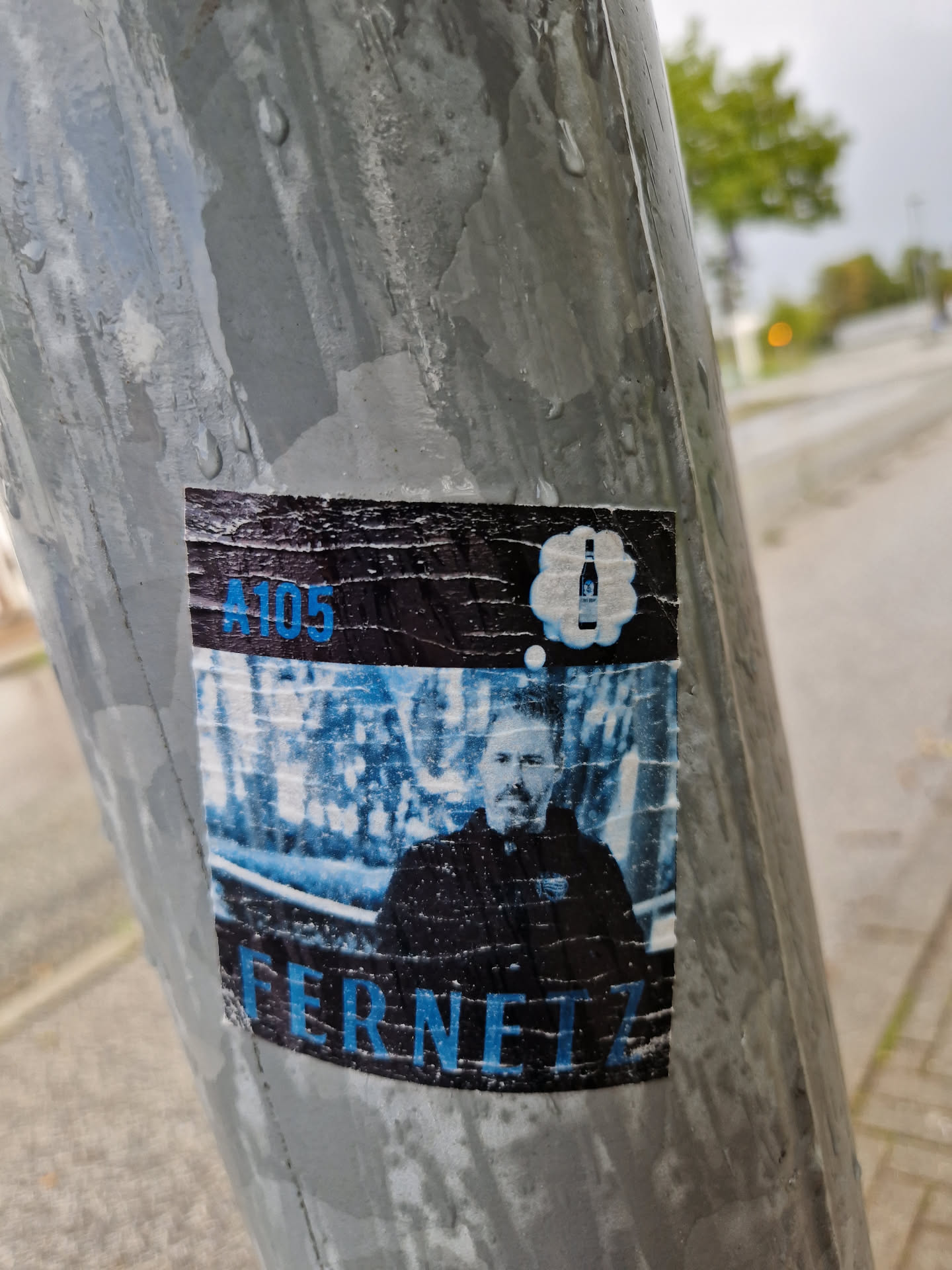 A sticker featuring a man's portrait with the text 'A105' and 'Fernet' underneath. A thought bubble with a bottle graphic is present above the man's head. The sticker shows some wear, suggesting it has been exposed to the elements for a while.