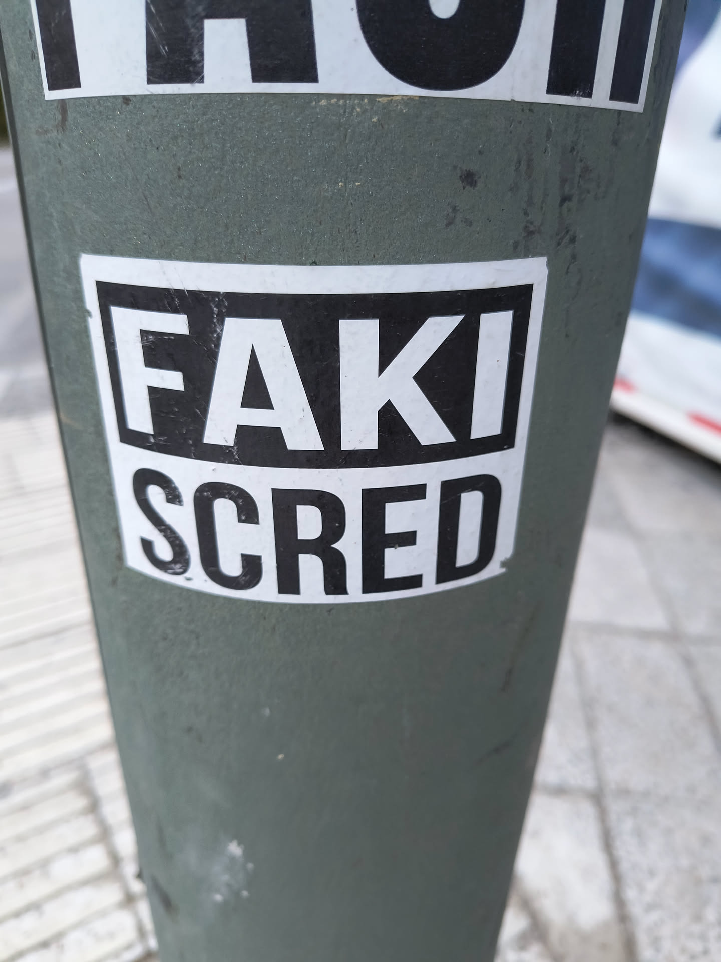 The sticker is rectangular with a white background and a thick black border. Centered on the sticker are two lines of text. The top line reads 'FAKI' in large, bold, sans-serif black capital letters. The bottom line reads 'SCRED' in similar bold, sans-serif black capital letters, slightly larger than the top line. The sticker appears to be applied to a dark green, cylindrical object, possibly a pole, which shows some scuff marks and texture.